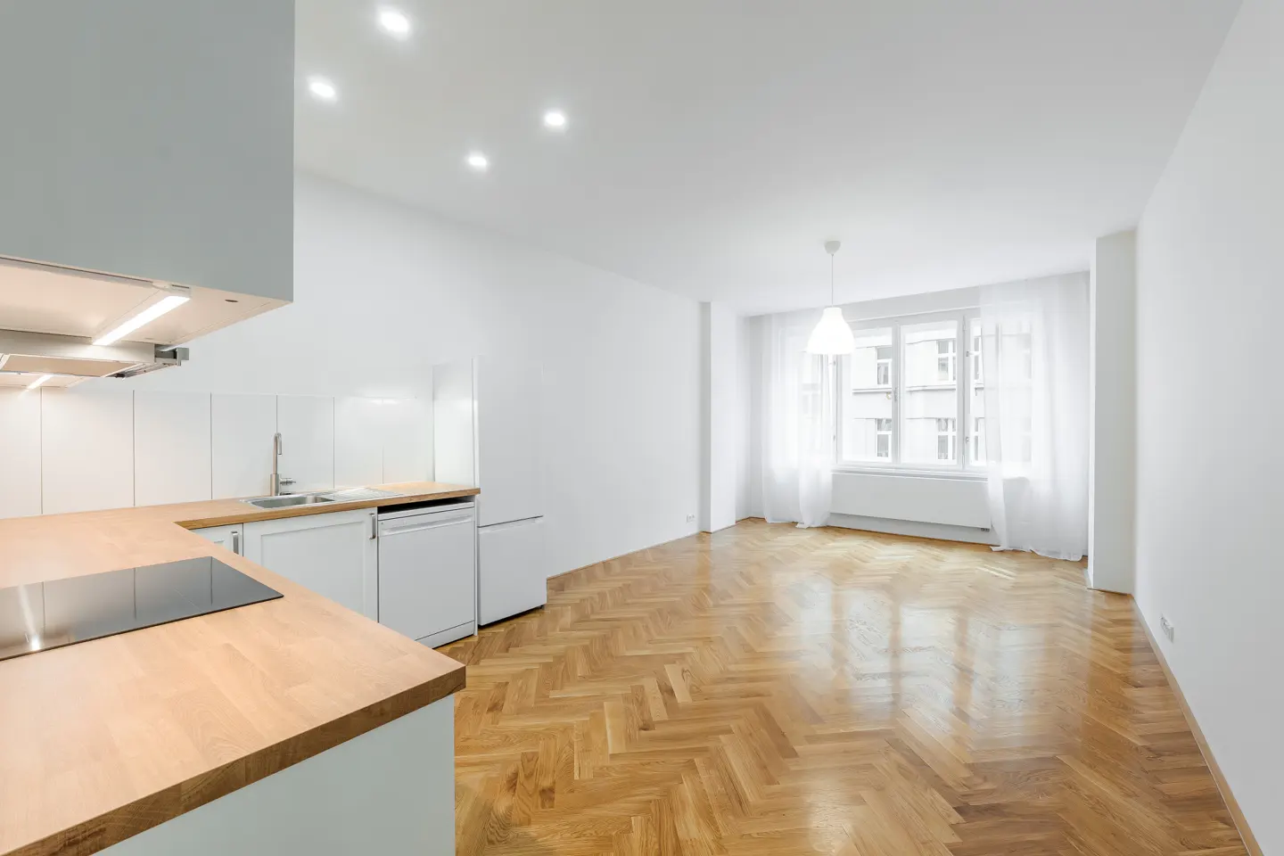 Bright, modern apartment with white walls, herringbone wood floors, and a kitchen with wood countertops. A large window with white curtains lets in natural light.