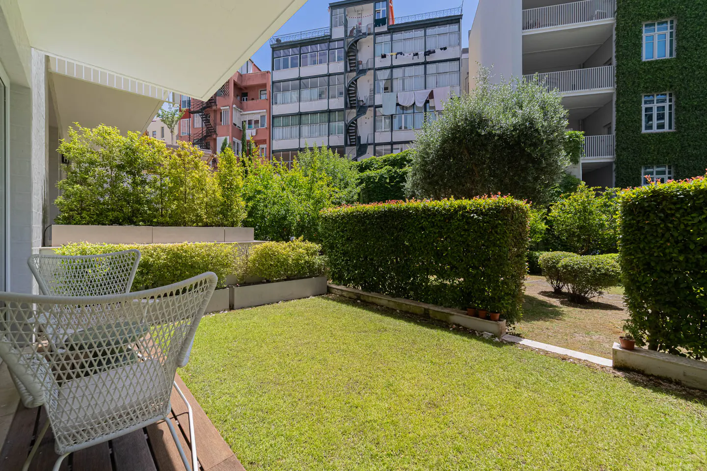 A patio with two white chairs overlooks a green lawn and manicured hedges, with buildings in the background.