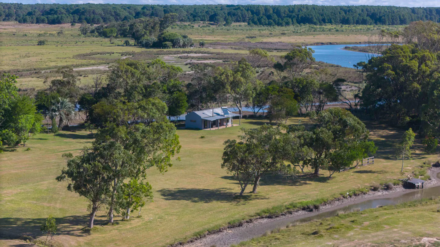 Aerial view of a gray house with a metal roof, surrounded by green trees and a river in a rural landscape.