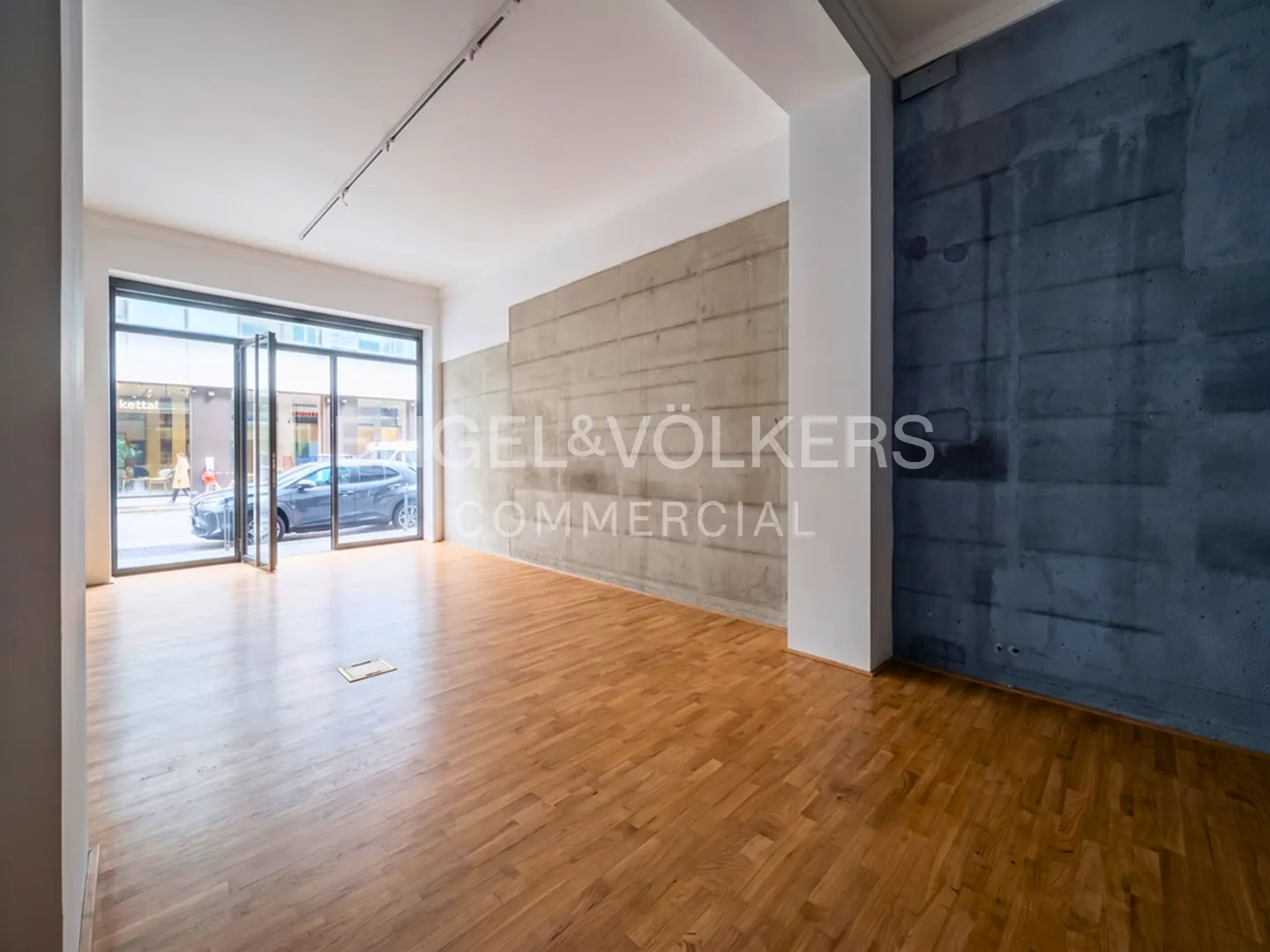Empty commercial space with wood floors, a light concrete wall, a dark blue accent wall, and large glass storefront windows.