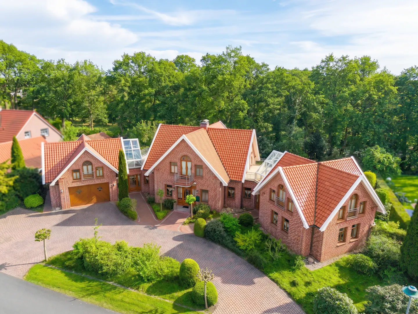 Aerial view of a large, red brick house with a red tile roof, surrounded by green trees and a paved driveway.