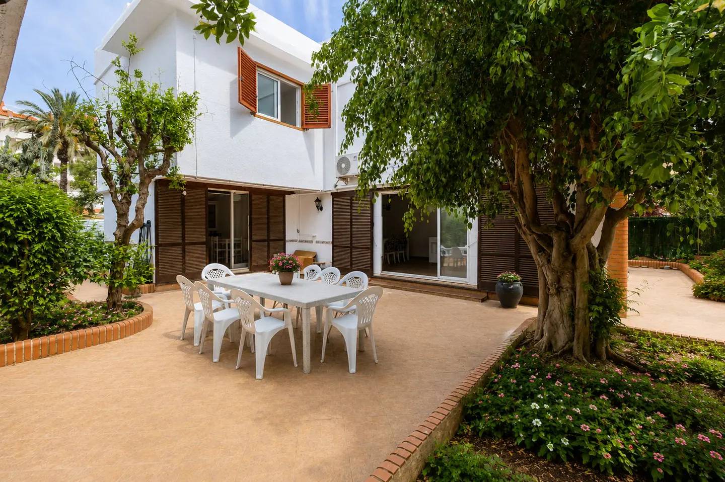A white house with a patio featuring a white table and chairs, surrounded by lush greenery.