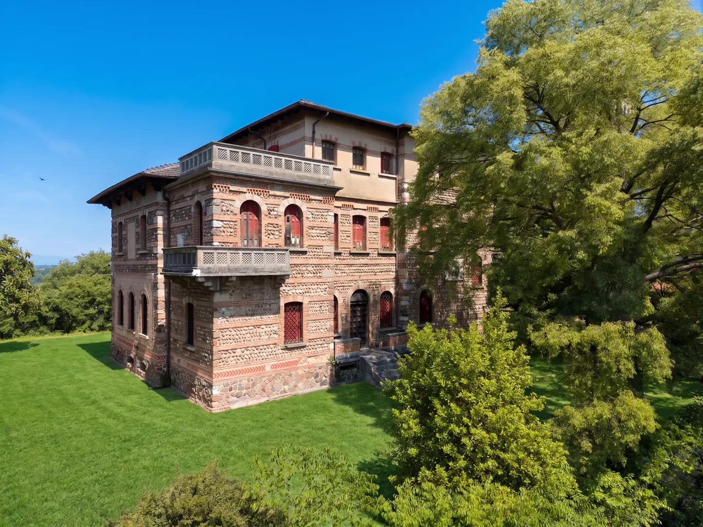A historic three-story brick villa with arched windows and a stone balcony, set on a lush green lawn under a blue sky.