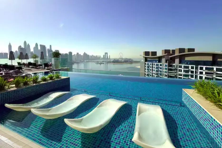 Rooftop infinity pool with white lounge chairs overlooking Dubai skyline and ocean on a sunny day.