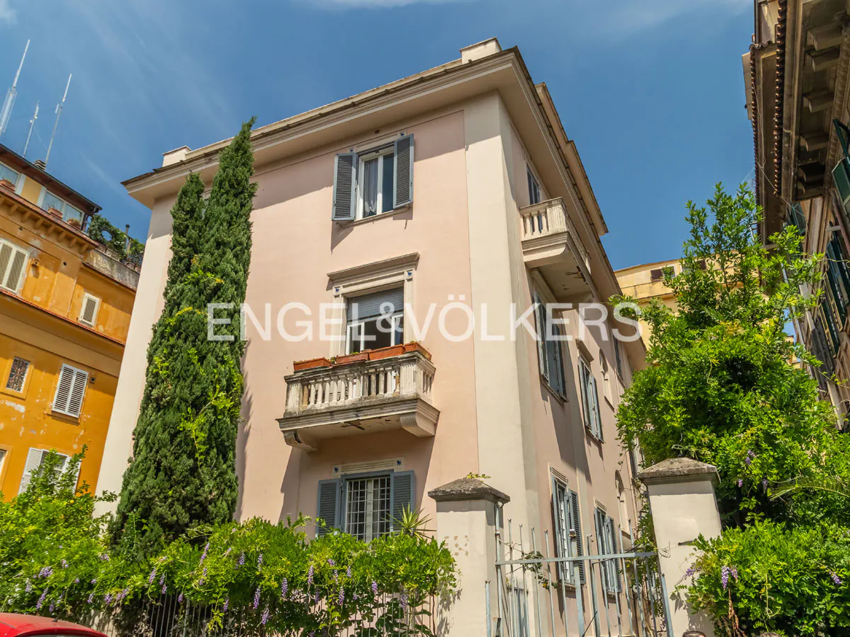 A pale pink multi-story building with a stone balcony, grey shutters, and a tall cypress tree against a bright blue sky.