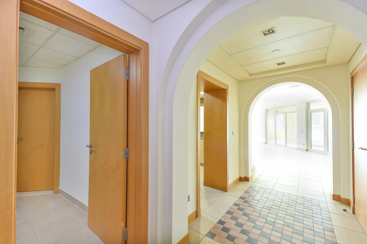 Interior view of a hallway with arched doorways and light wood doors. Tiled floor with a checkered pattern. Cream-colored walls and ceiling.