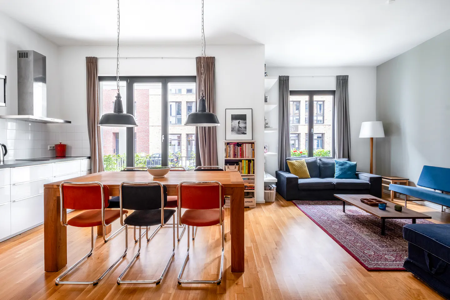 Open-plan living area with a wooden dining table, colorful chairs, a dark sofa, and a patterned rug on hardwood floors.