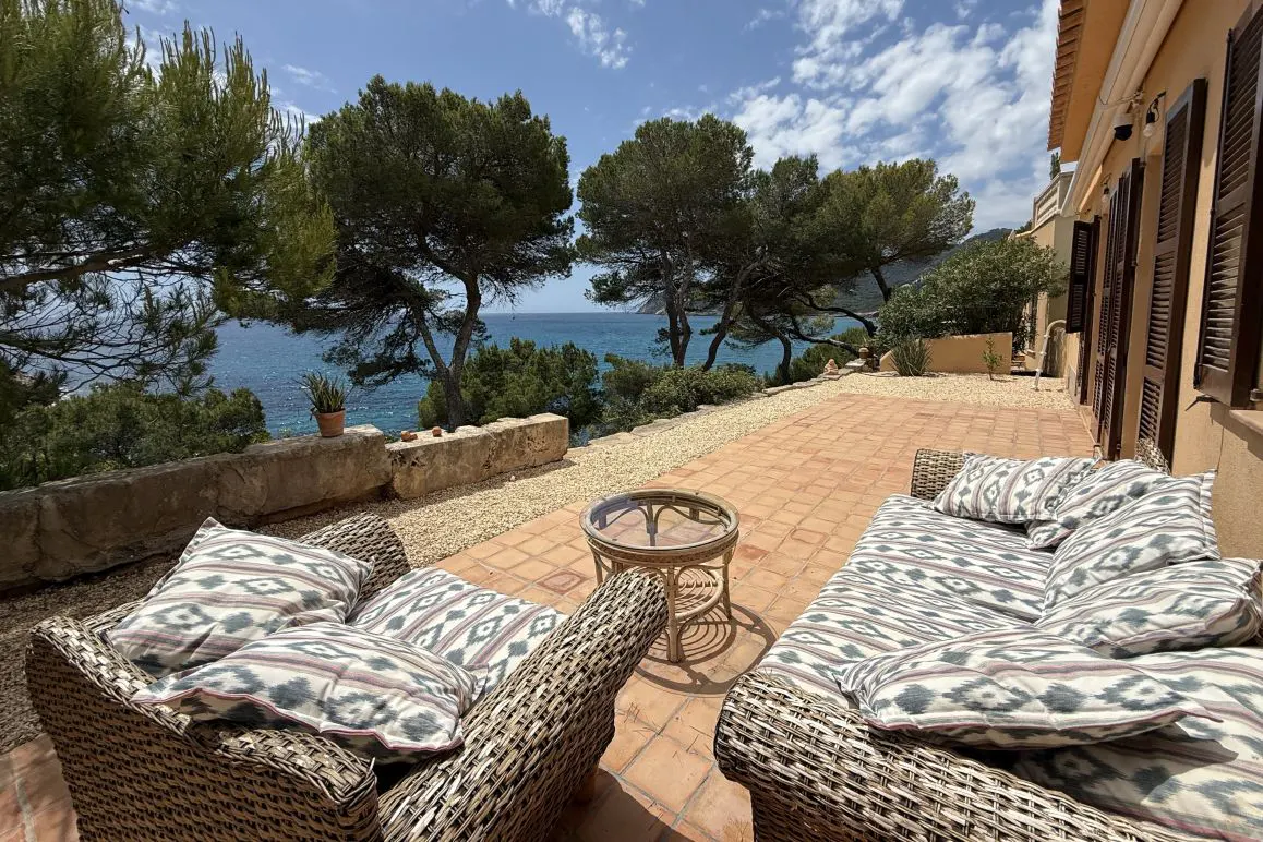 Outdoor patio with wicker furniture, patterned cushions, and a glass table overlooking the ocean and trees.