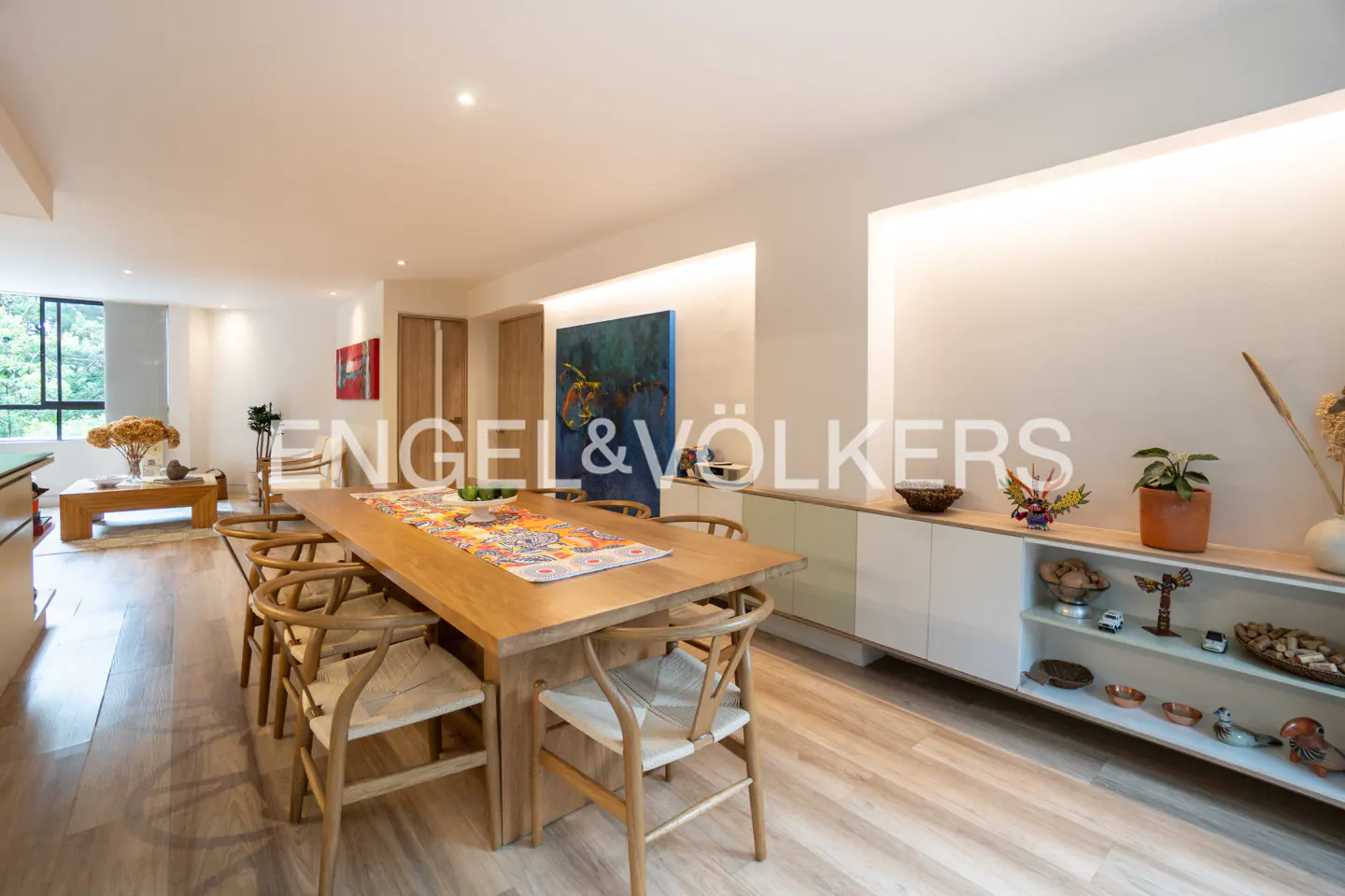 Bright dining room with a long wooden table, chairs, and colorful runner. White cabinets line the wall with decorative items. Engel & Völkers logo visible.