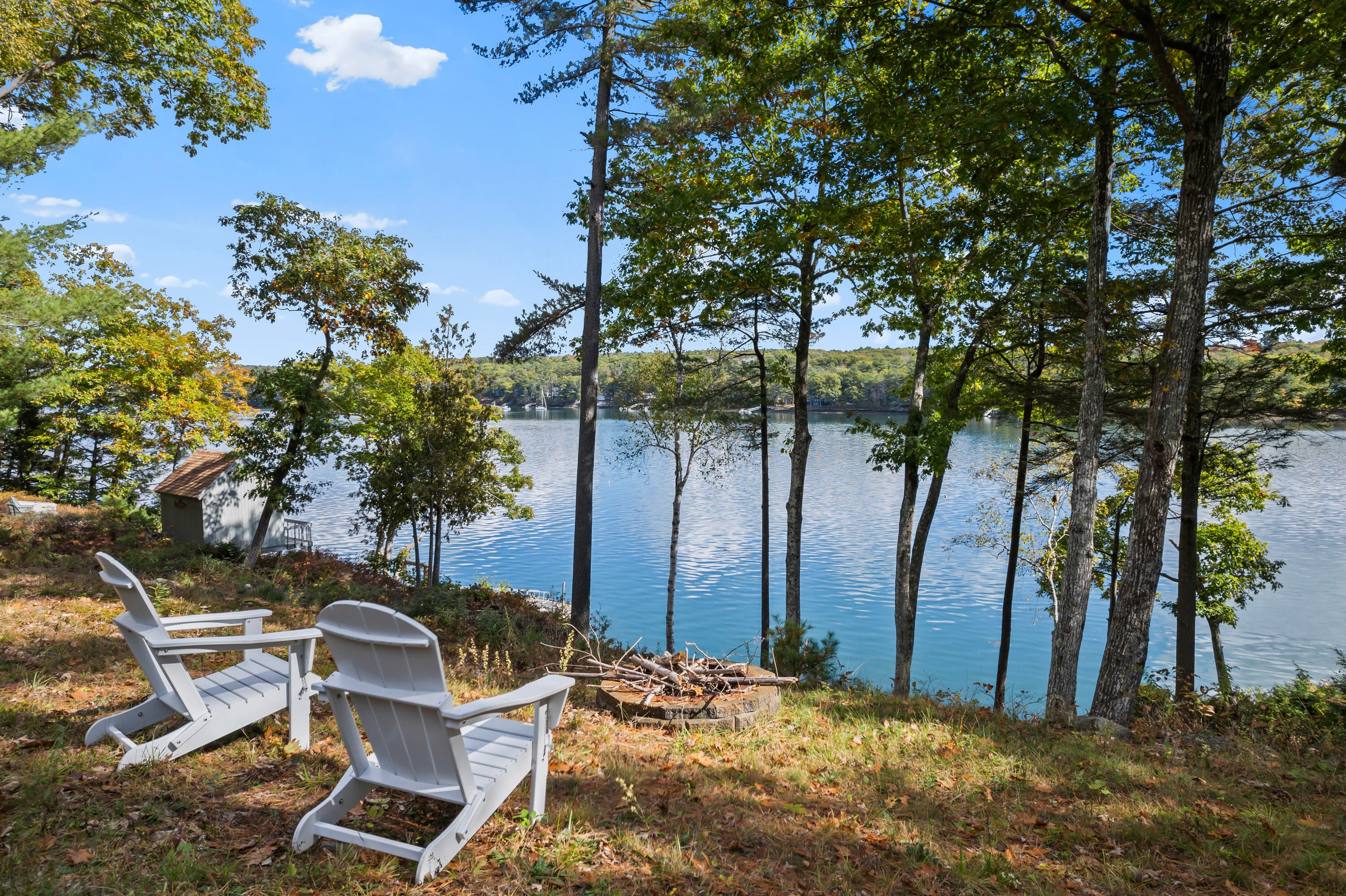 Two white Adirondack chairs face a stone fire pit overlooking a calm lake surrounded by lush green trees. [AI-generated text]