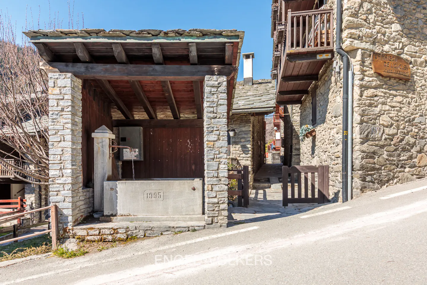 Stone water fountain with a wooden roof, dated 1995, stands on a street corner in a European village with stone buildings.
