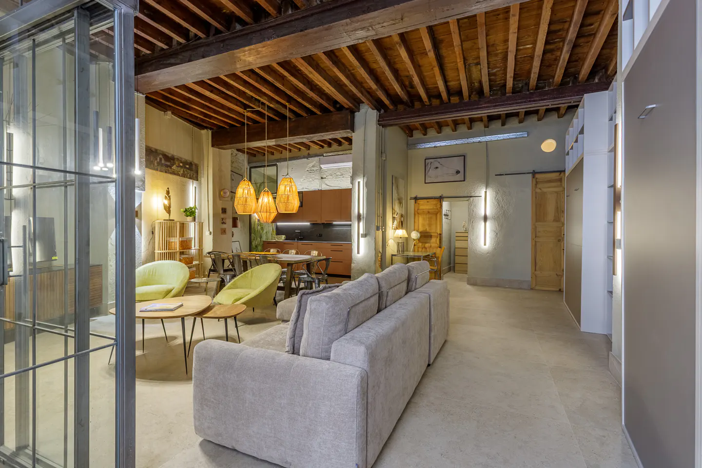 Interior view of a living room with a gray sofa, wooden ceiling beams, and woven pendant lights. A dining table and kitchen are in the background.