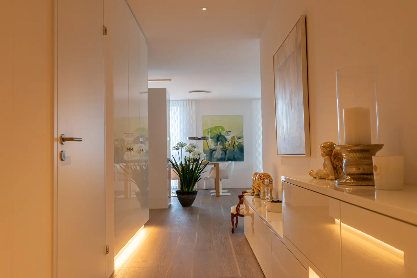 Hallway view of a modern home with white walls, light wood floors, and recessed lighting. A painting and decor adorn a white console table.