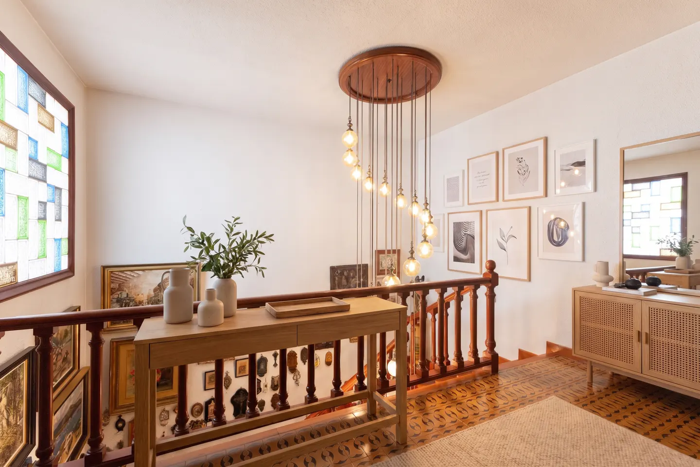 Bright landing with wood railing, patterned tile floor, and gallery wall. A modern chandelier hangs above a console table with vases.