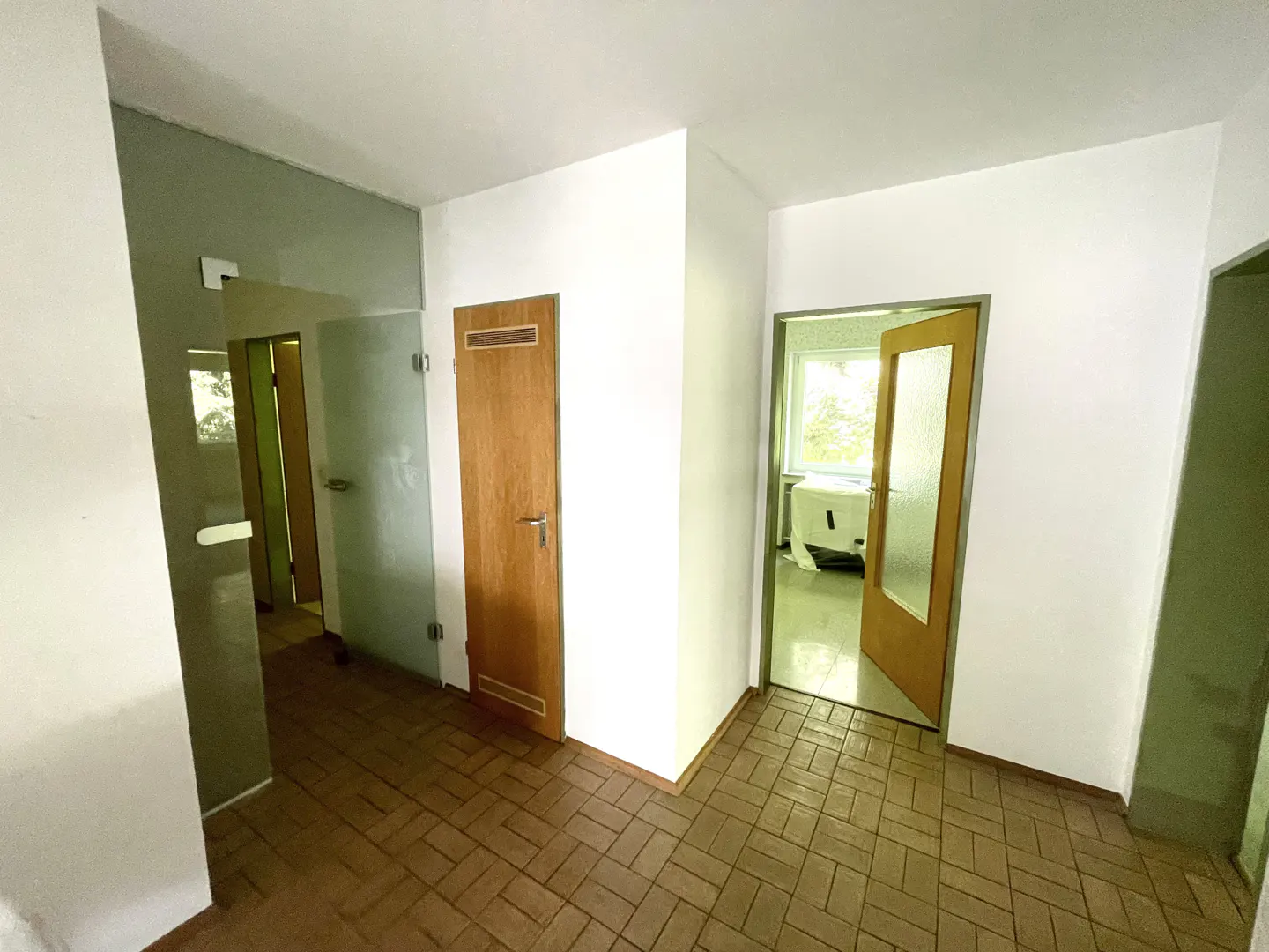 Hallway with brown tiled floor, white walls, a wooden door, and a glass door leading to a room with a window.