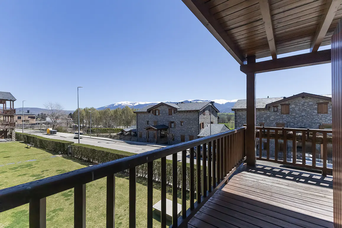 View from a wooden balcony overlooking stone houses, a green lawn, and snow-capped mountains under a clear blue sky.