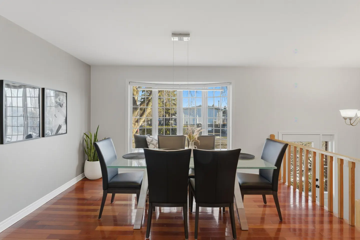 Bright dining room with a glass table, black chairs, and hardwood floors. A large window overlooks a yard with trees.