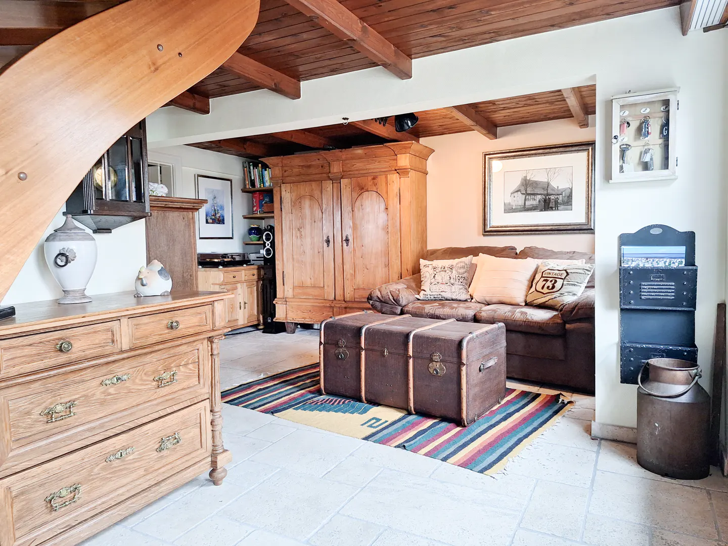 Living room with wood-beamed ceiling, brown sofa, and trunk-style coffee table on a colorful rug. A wooden dresser is on the left.