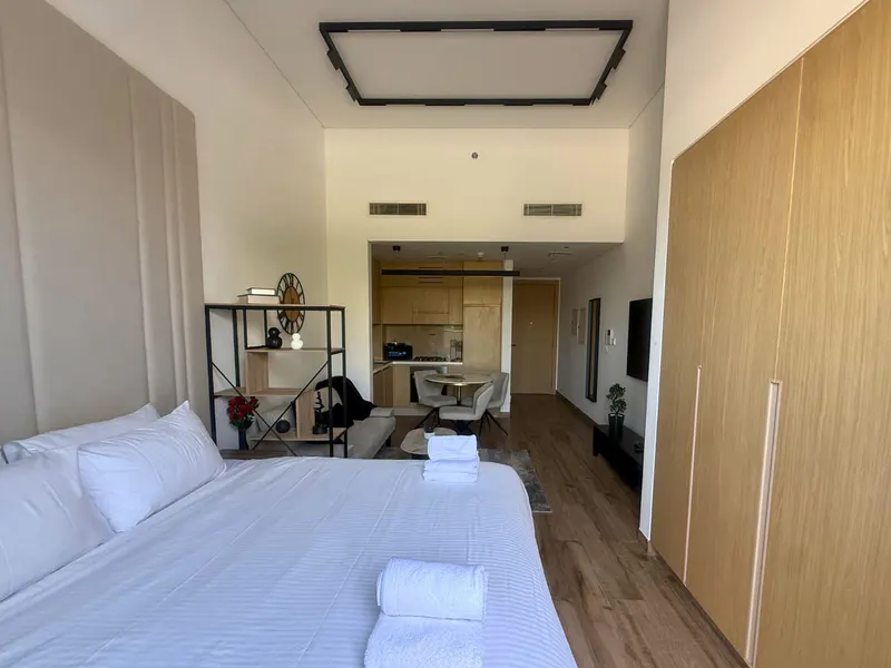 Bright studio apartment with a white bed, open kitchen, dining area, and wooden floors. A black shelf and large wooden closet add storage.