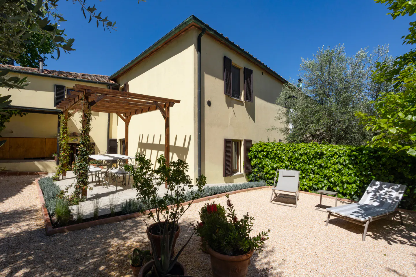 Exterior view of a yellow house with brown shutters, a pergola, and outdoor seating on a gravel patio.