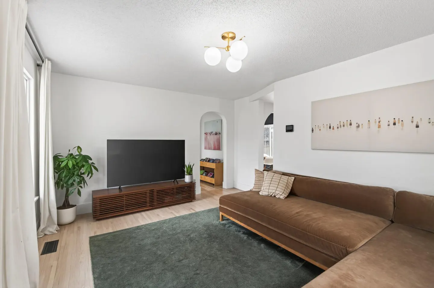 Living room with a brown sectional sofa, a TV on a wooden stand, and a green rug. White walls and light wood floors.