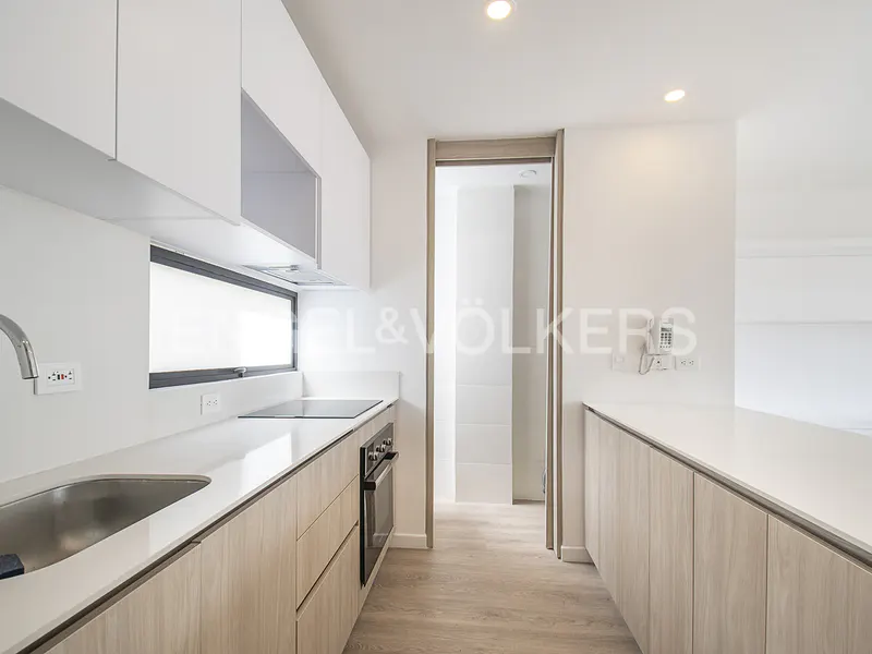 Modern kitchen with white upper cabinets, light wood lower cabinets, white countertops, and a doorway to a hallway.
