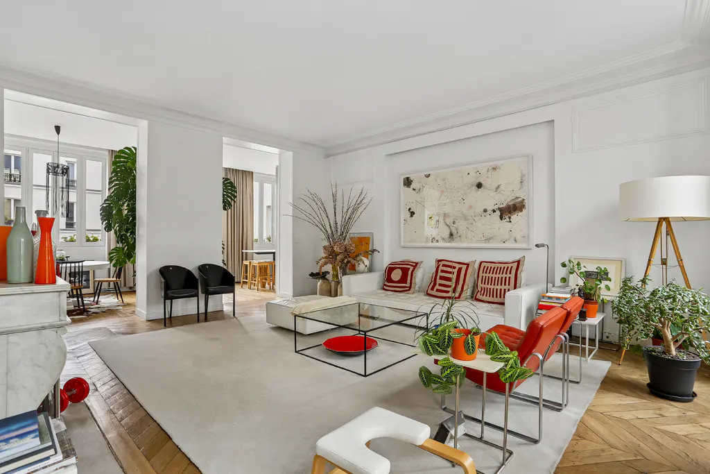 Bright, airy living room with white walls, wood floors, and a large white rug. A white sofa with red pillows sits near a glass coffee table. Plants and art add pops of color.