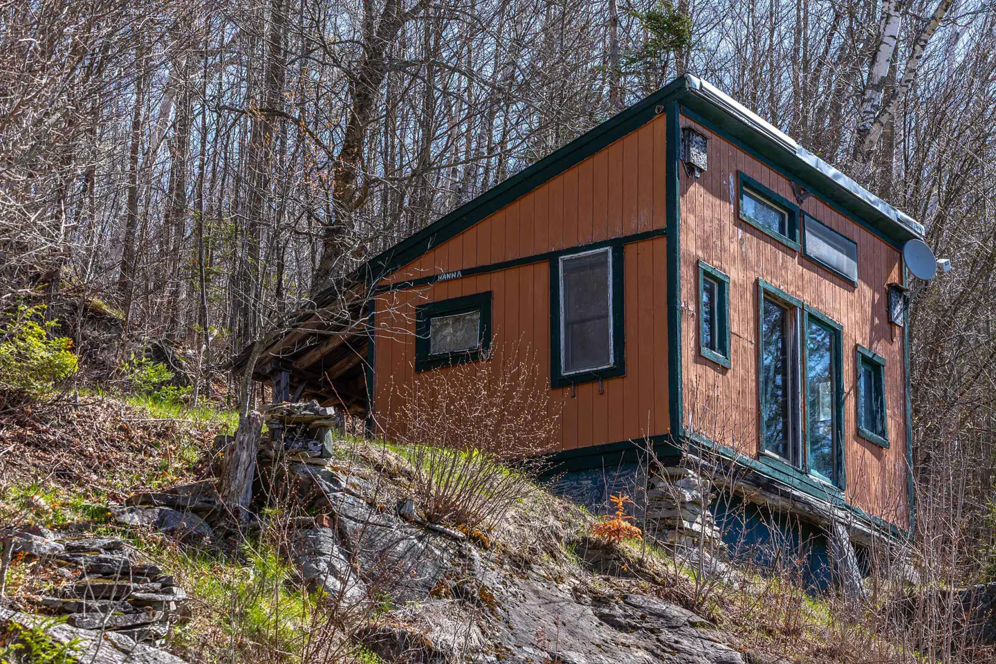 Rustic brown cabin with green trim, nestled on a rocky hillside surrounded by bare trees. Windows reflect the sky.