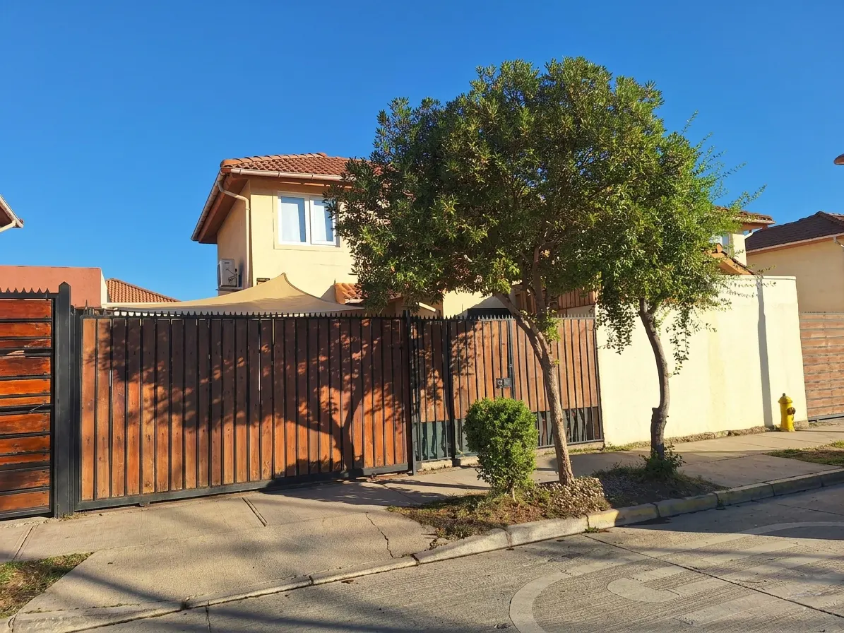 Two-story tan house with a brown wood gate and green trees under a clear blue sky.