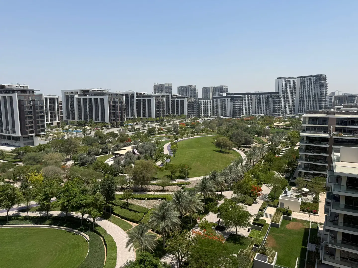 Aerial view of modern apartment buildings surrounding a green park with trees and walking paths on a sunny day.