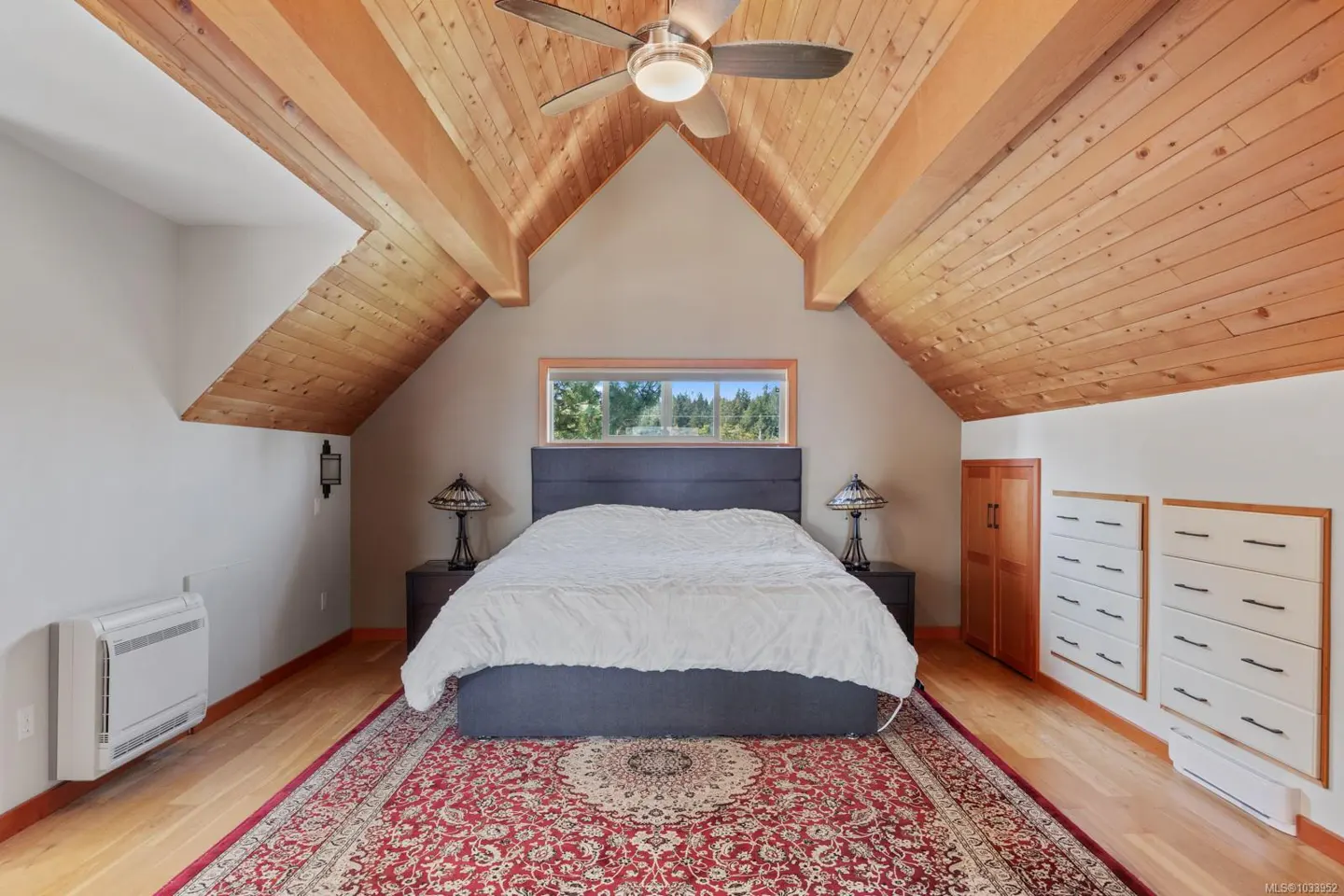 Bedroom with wood-paneled ceiling, red rug, and bed with a gray headboard. A window is above the bed.