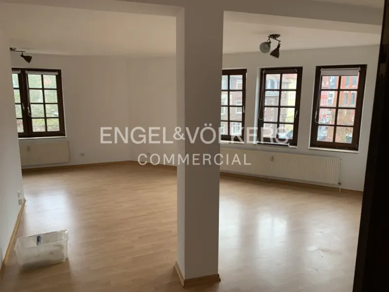 Bright, empty room with light wood floors, white walls, and dark-framed windows. A support column divides the space. Engel & Völkers logo is visible.