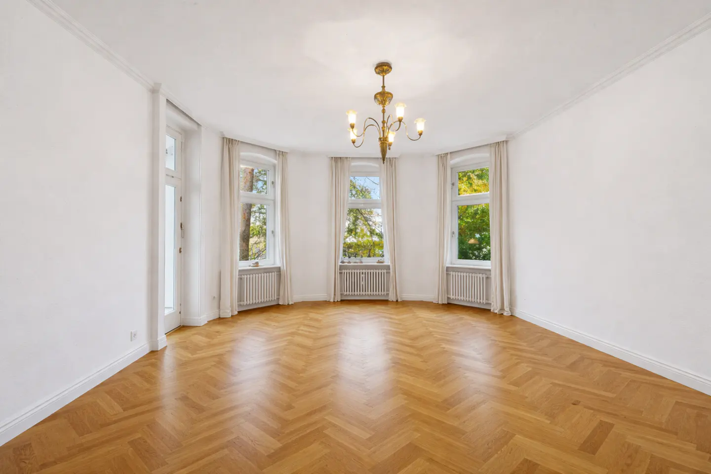 Bright, empty room with herringbone wood floors, white walls, and a gold chandelier. Three windows with white curtains overlook trees.