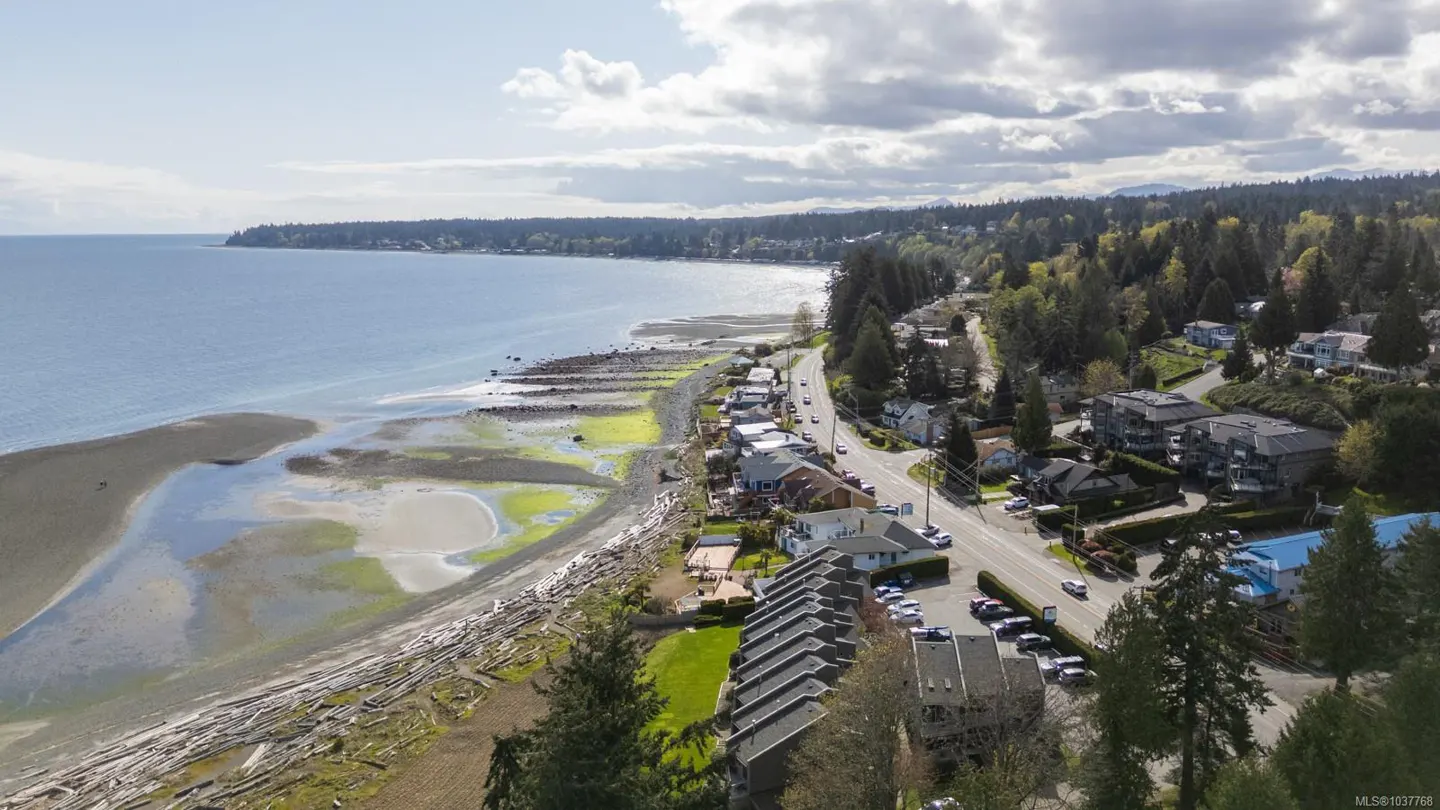 Aerial view of a coastal community with a sandy beach, blue ocean, and green trees. Houses and a road run along the coastline. Cloudy sky above.