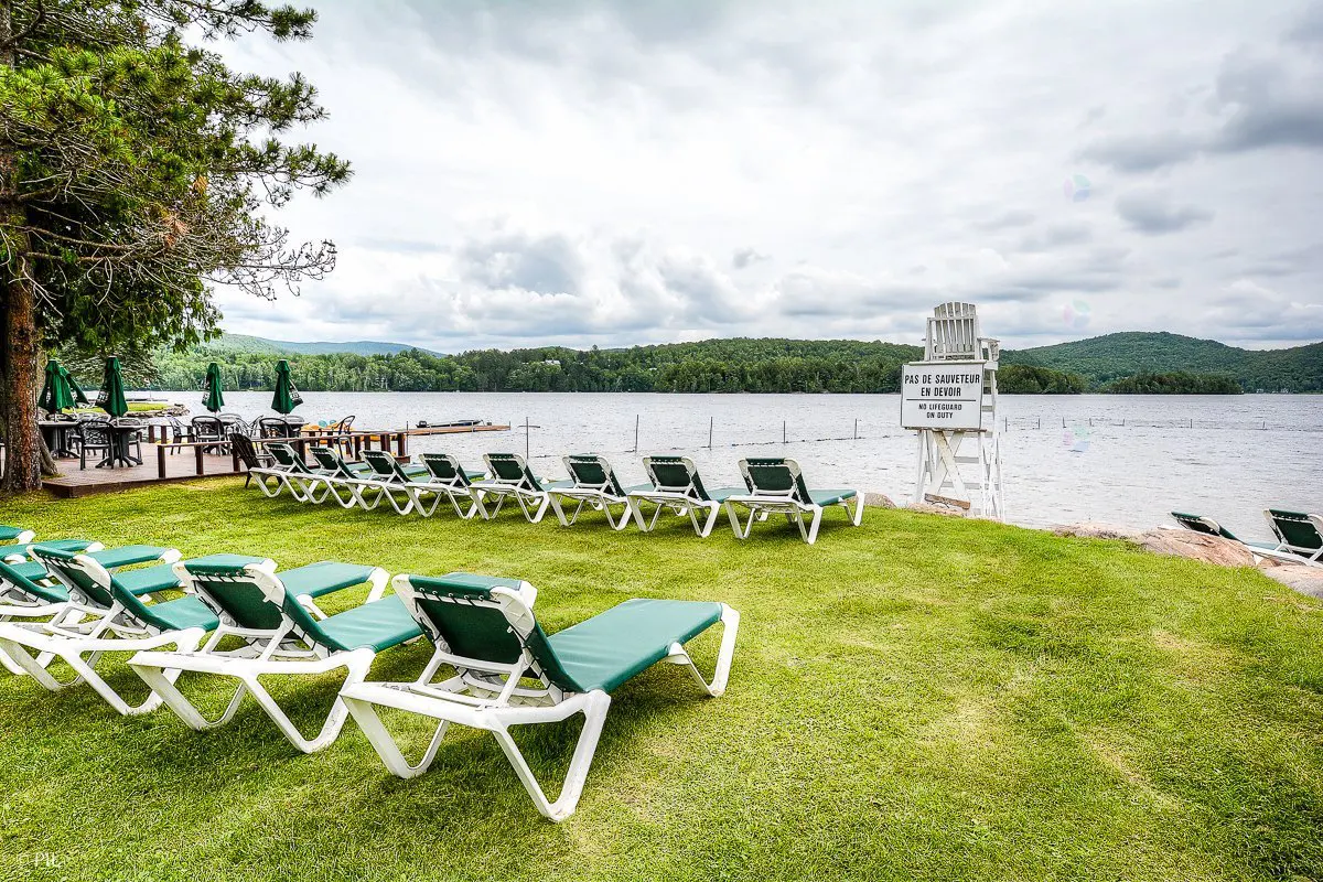 Lounge chairs on a grassy lawn overlook a lake with mountains in the distance. A lifeguard stand is visible near the water.