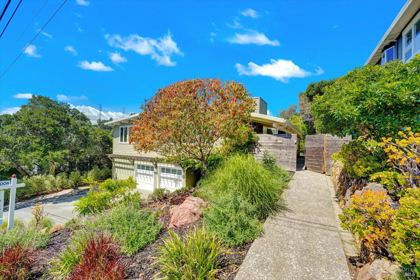 Two-story light green house with a two-car garage, a tree with red leaves, and a concrete walkway. Blue sky with clouds.