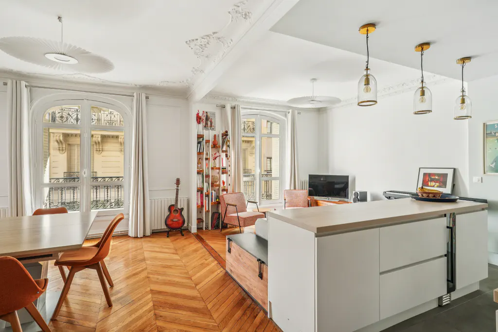 Bright, airy apartment interior with herringbone wood floors, white walls, and large windows overlooking a Parisian street.