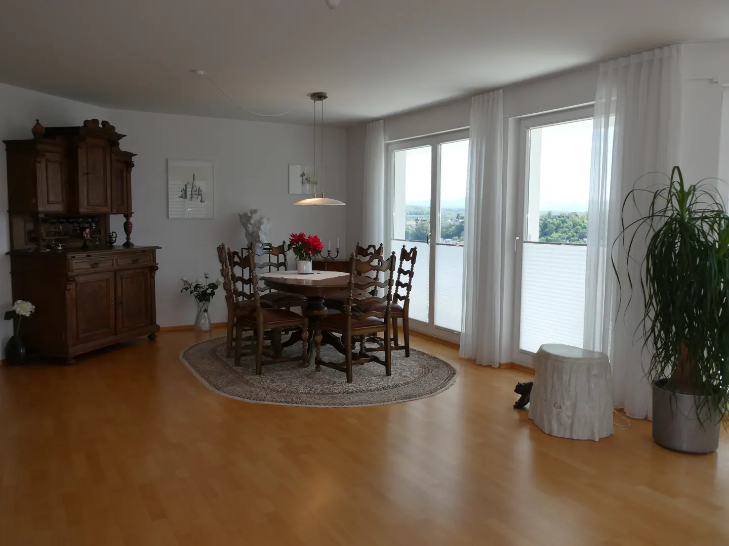 A dining room with wooden floors, a table with chairs, and a large wooden cabinet. White curtains frame the windows.