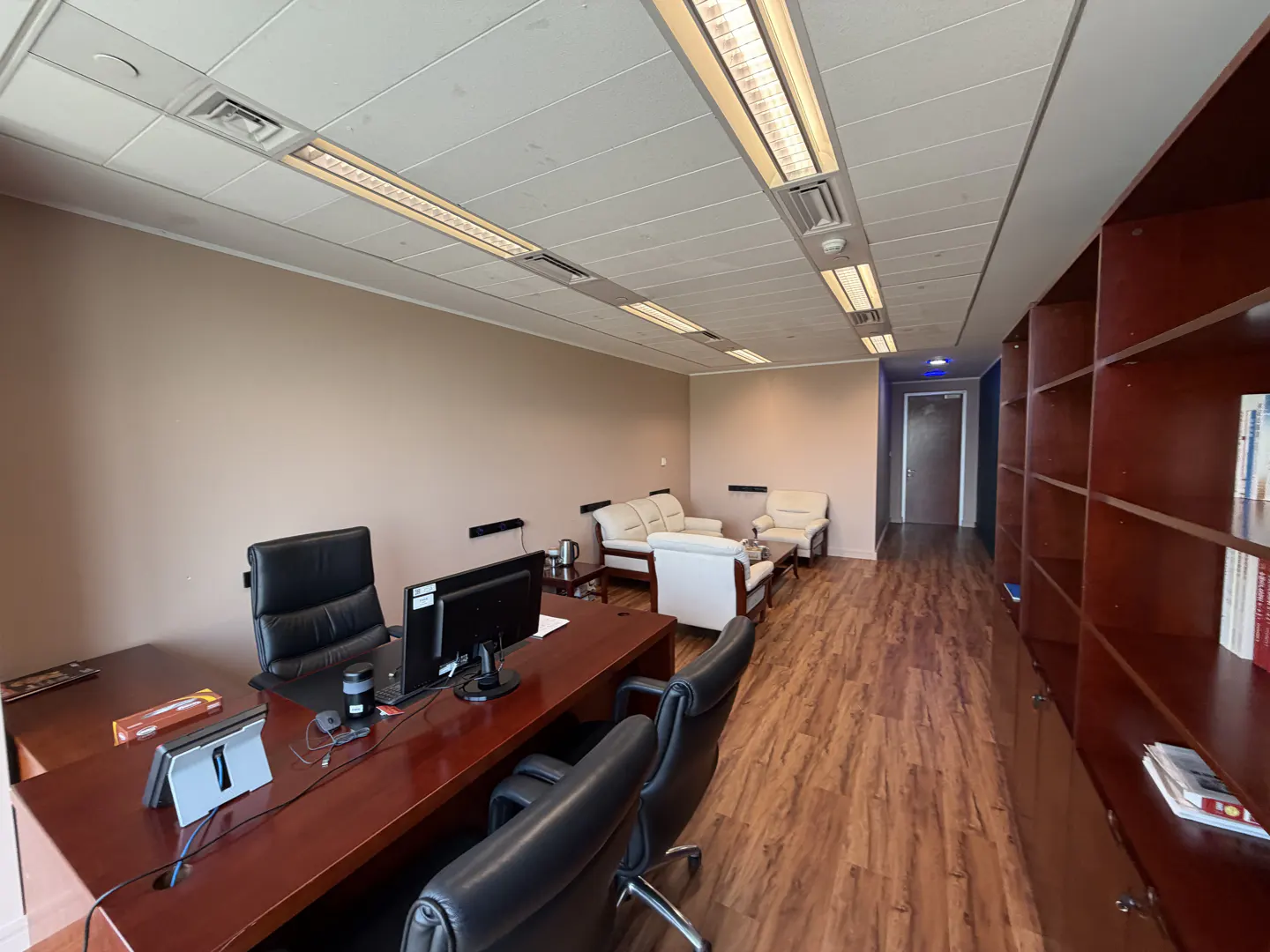 Office interior with a large wooden desk, black chairs, and a seating area with white sofas. A tall bookshelf lines the right wall.