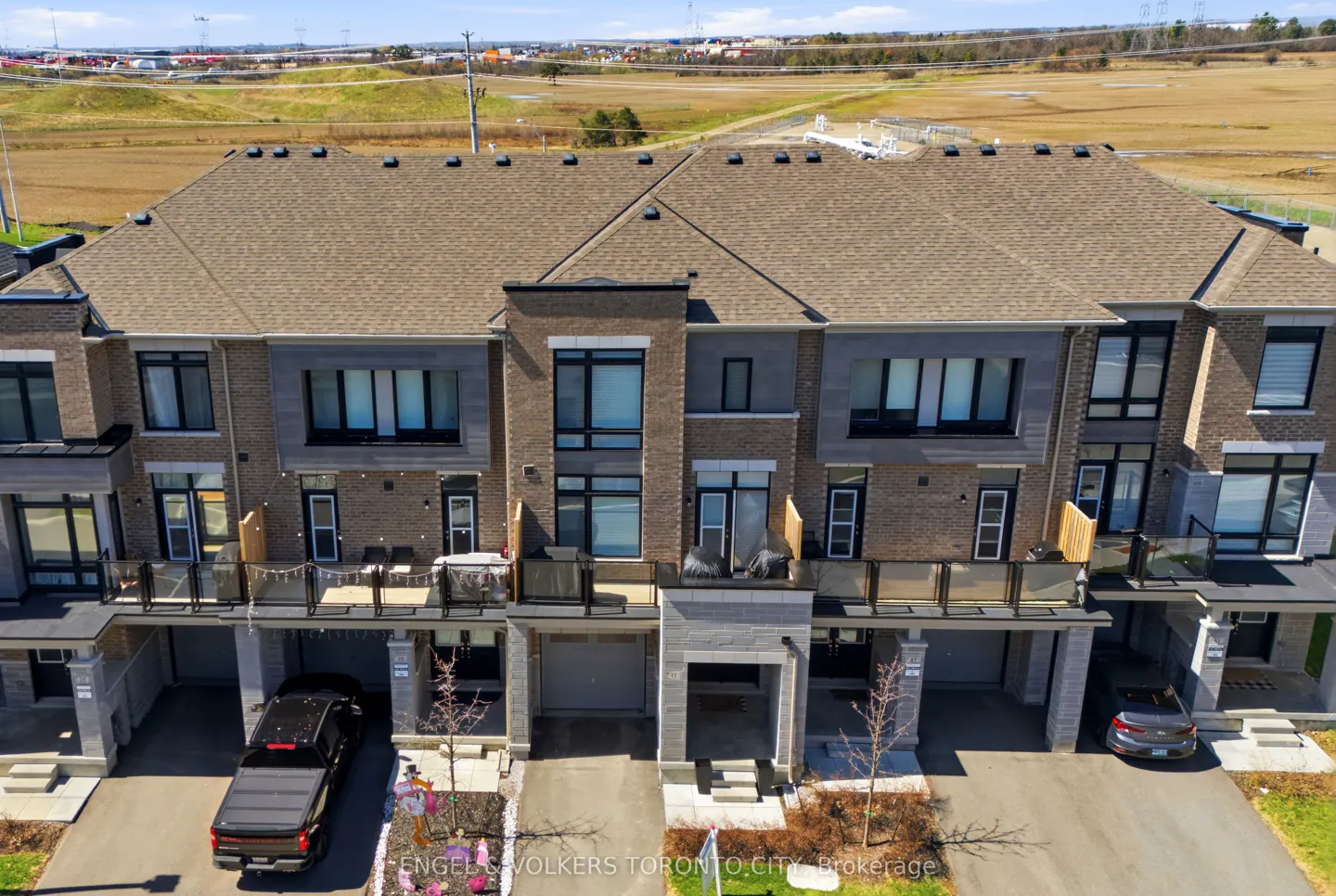 Aerial view of modern, multi-story townhouses with brown brick, black trim, and attached garages. Cars are parked in driveways.