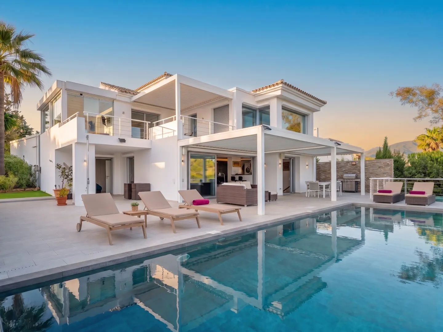 Modern white villa with a pool, lounge chairs, and outdoor dining area under a pergola against a blue sky.