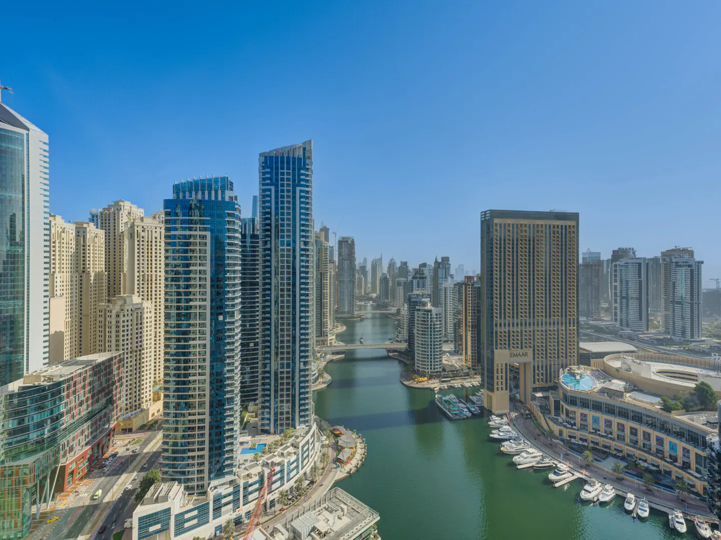 Aerial view of Dubai Marina with skyscrapers, a canal, boats, and a clear blue sky.