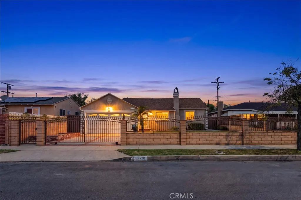 A single-story home with a brown roof and beige exterior, enclosed by a brick wall and black iron gate at dusk.