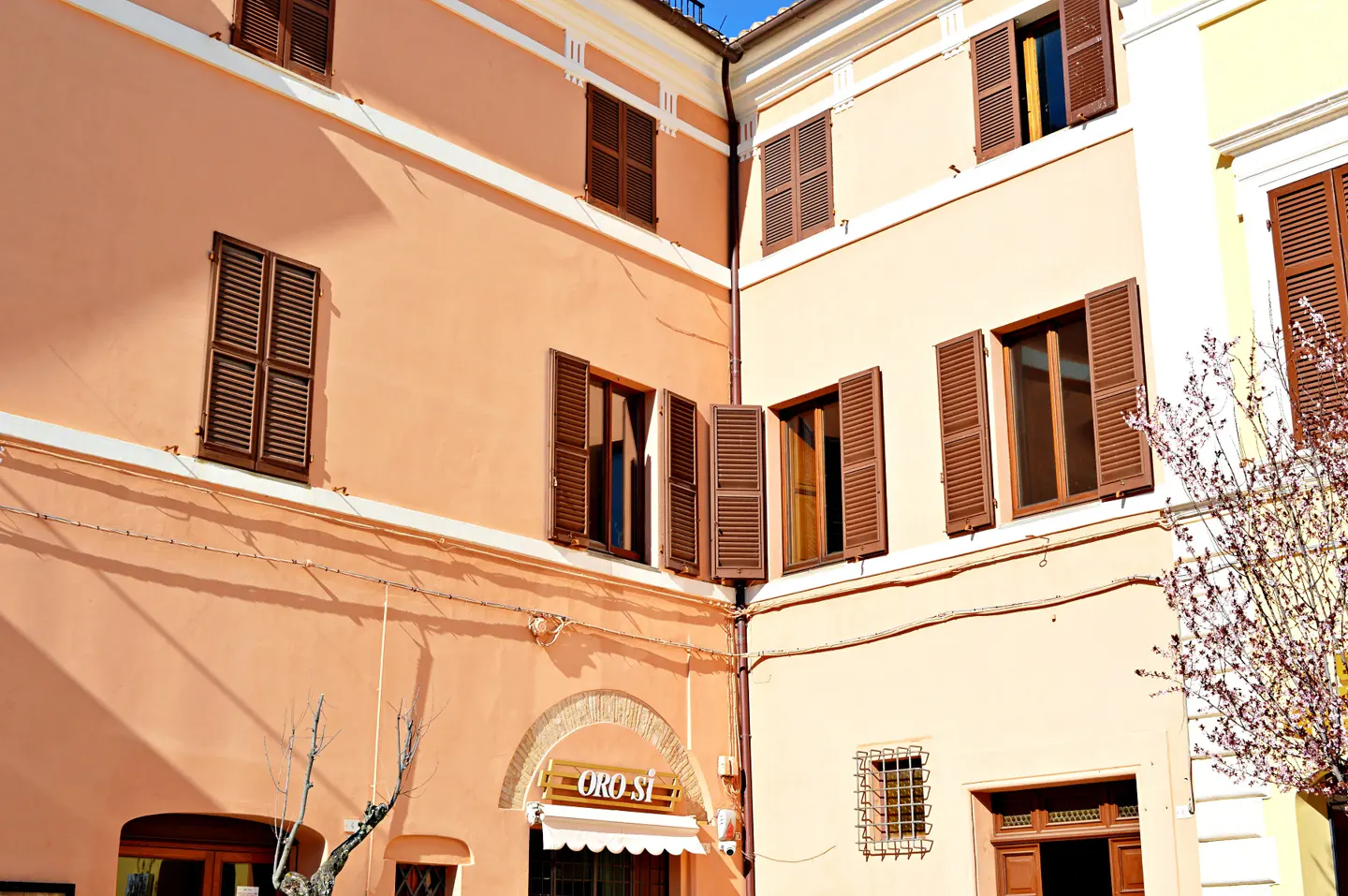 Exterior view of peach and cream buildings with brown shutters, some open, and a shop sign reading "ORO SI" under an arched doorway.