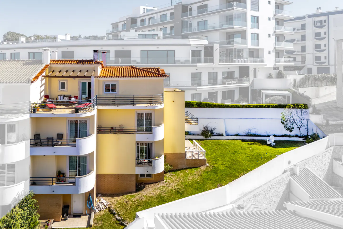 Exterior view of a yellow apartment building with balconies and a red tile roof, overlooking a green lawn and white wall.