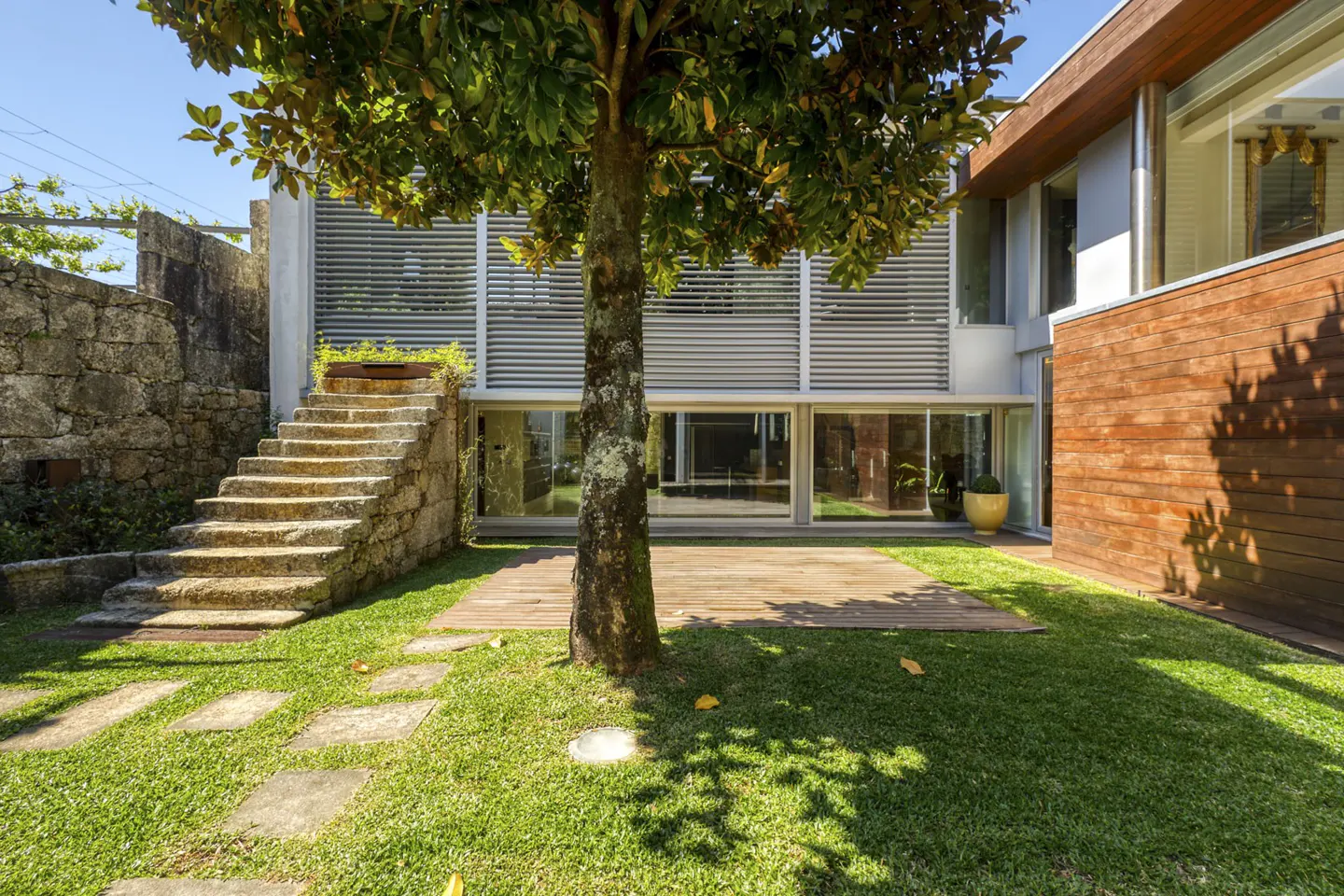 Modern home exterior with a grassy yard, stone stairs, a central tree, and a wooden deck under a bright blue sky.