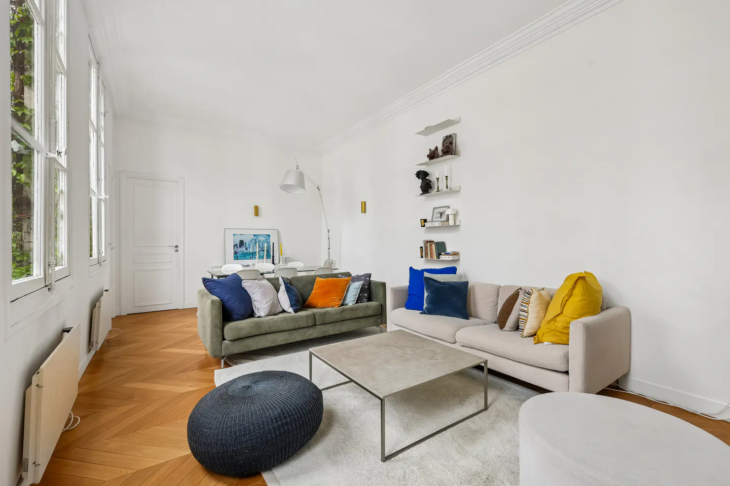 Bright living room with parquet floors, white walls, and large windows. Two sofas face each other over a light gray rug and coffee table.