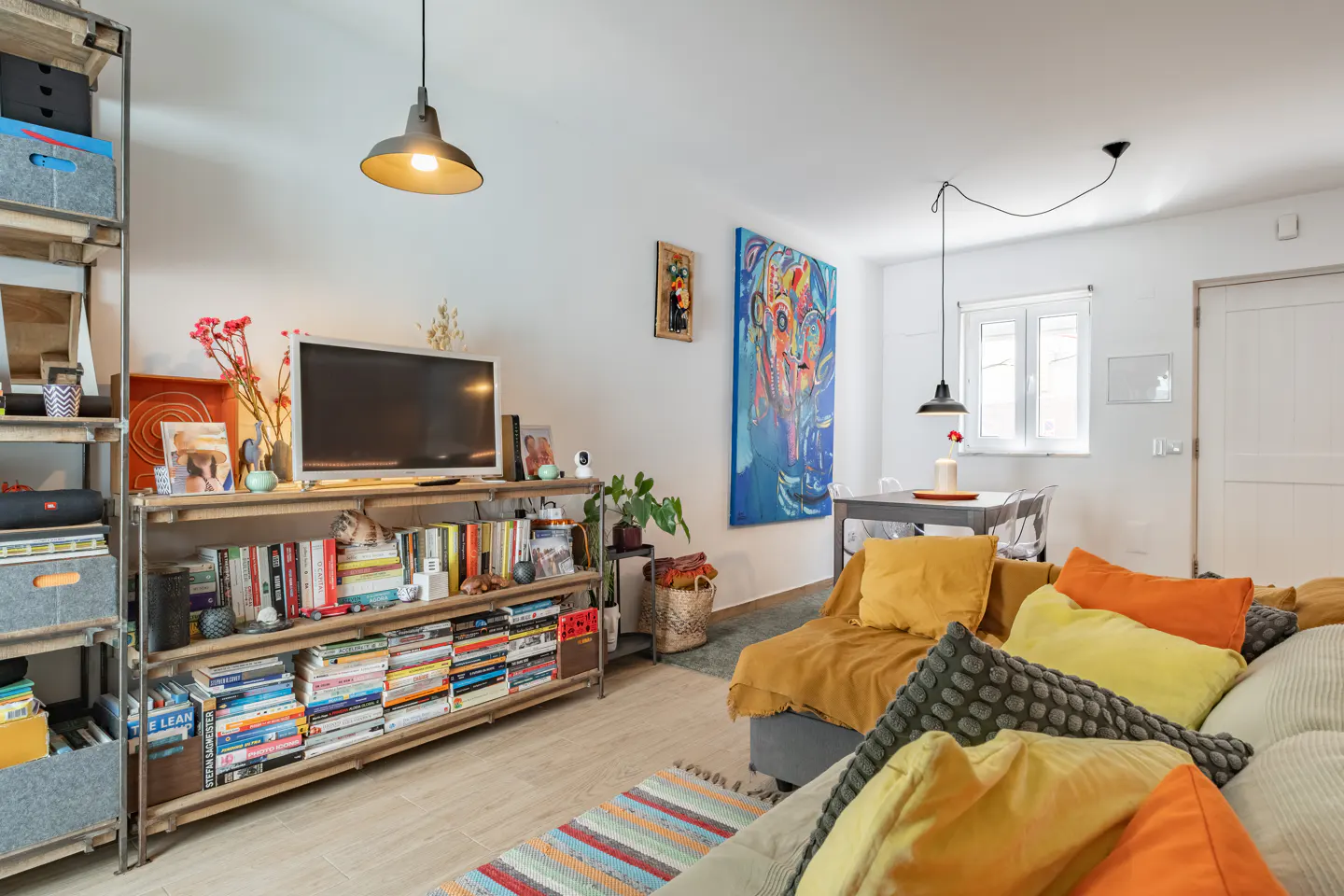 Bright living room with a bookshelf filled with books, a TV, and colorful decor. A large blue painting hangs on the wall. A sofa with yellow and orange pillows.