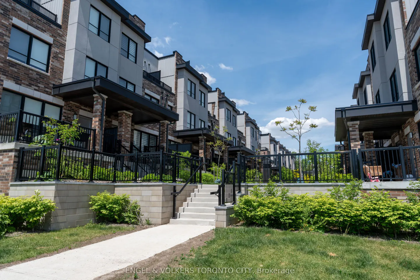 Modern brick and grey townhouses with black railings, concrete stairs, and green landscaping under a bright blue sky.