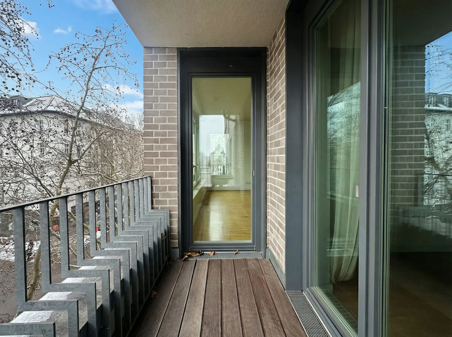 Modern balcony with wooden decking, gray brick walls, metal railing, and a glass door looking into a bright room.